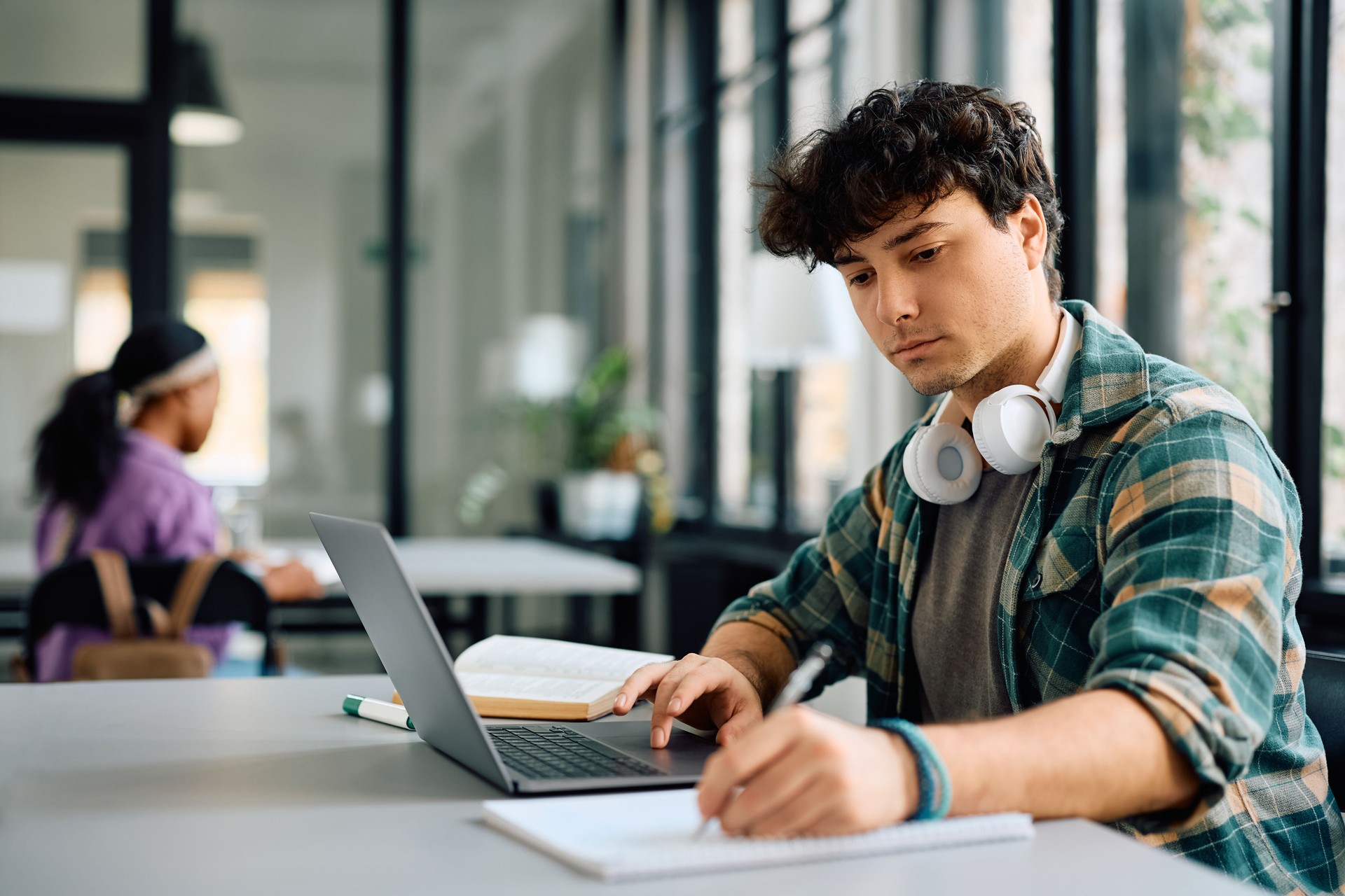 Jeune étudiant prenant des notes tout en apprenant en ligne sur un ordinateur portable à l’université.